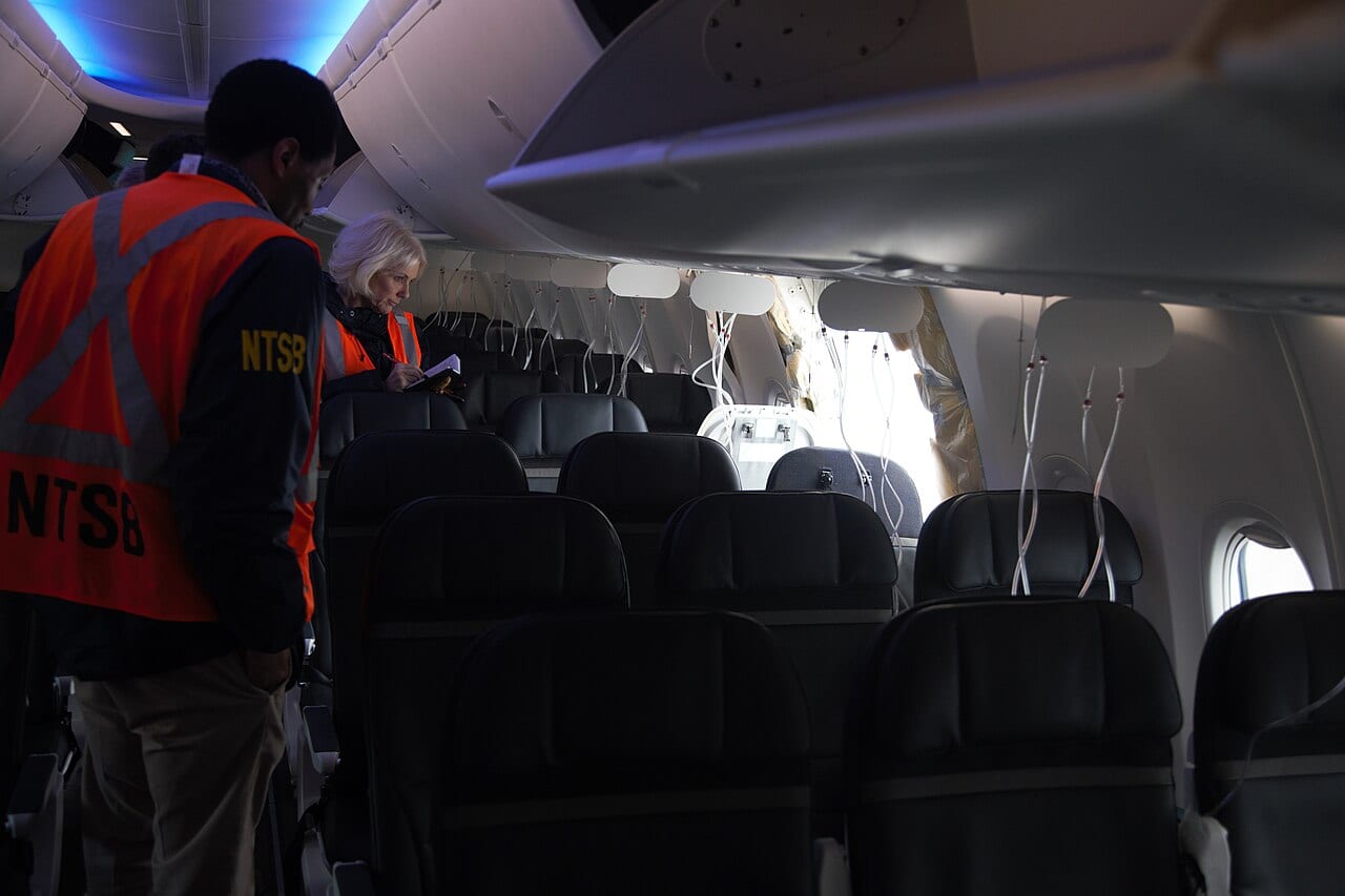 Interior of the Flight 1282 cabin showing the rectangular gap left by the missing door plug, insulation and wiring exposed, daylight visible through the hole.