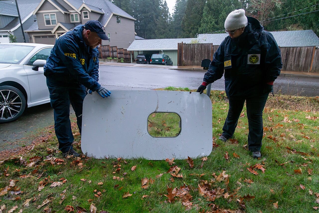 Two NTSB investigators lifting the recovered door plug panel from a residential lawn, its window cut-out visible, the morning after it was found.