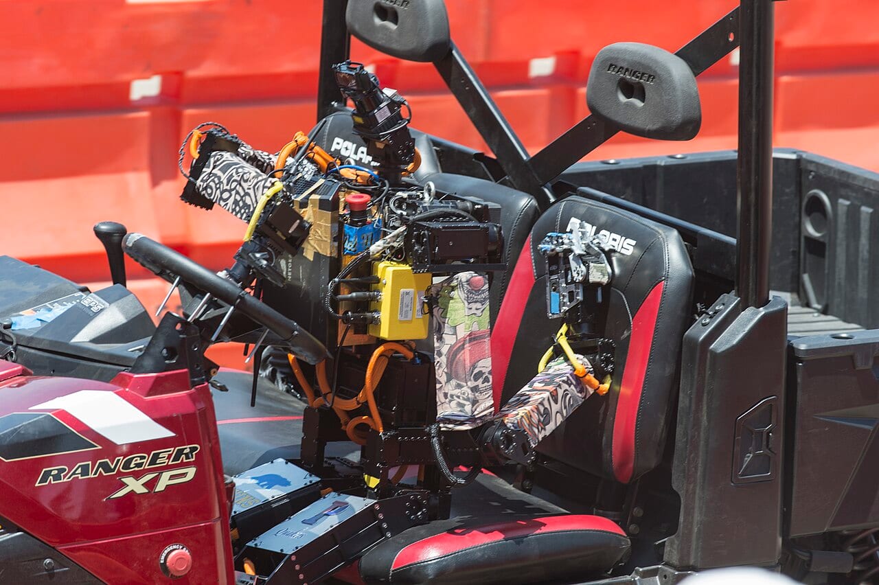 The humanoid robot THOR manipulating vehicle controls during the DARPA Robotics Challenge, its hands and arms positioned around the equipment.