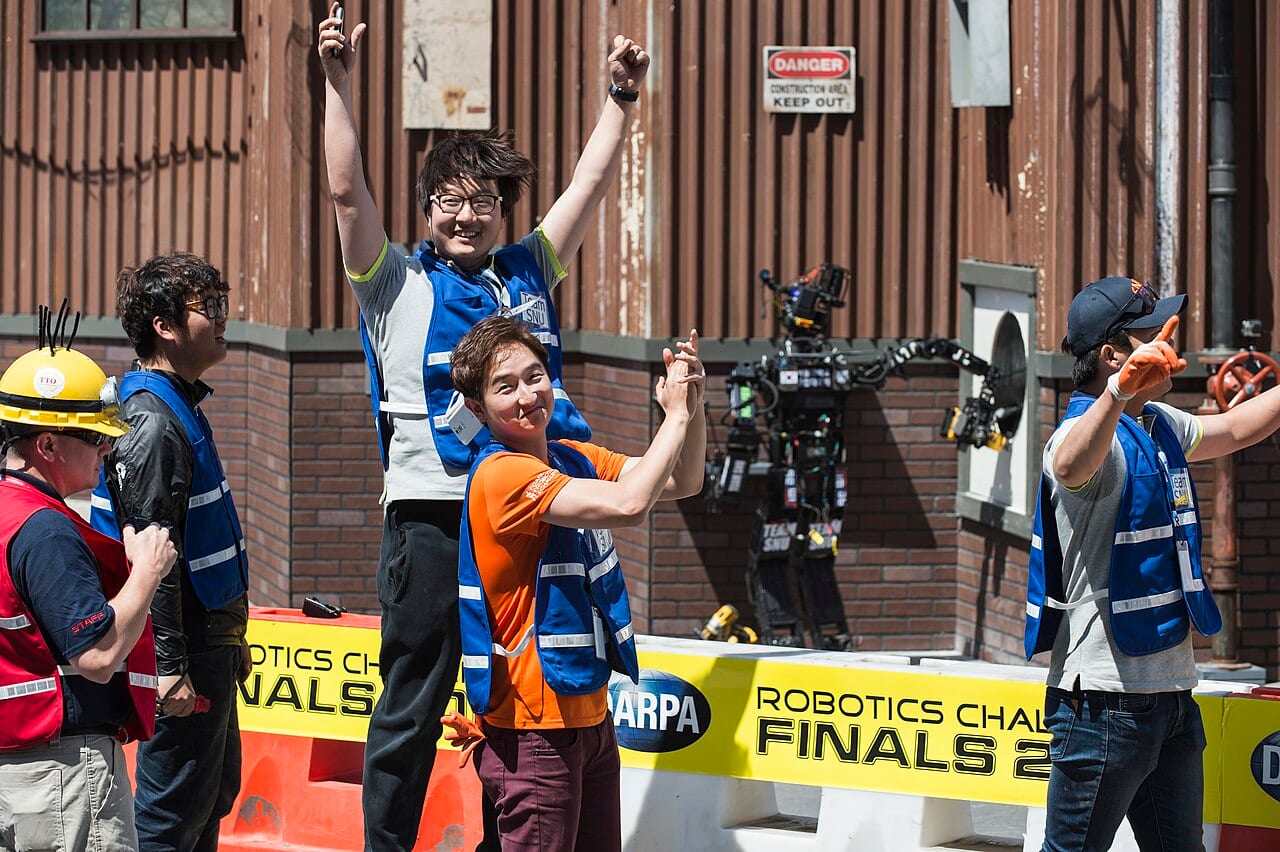 The humanoid robot THORMANG, entered by Seoul National University, completing a task during the DARPA Robotics Challenge as its team celebrates.