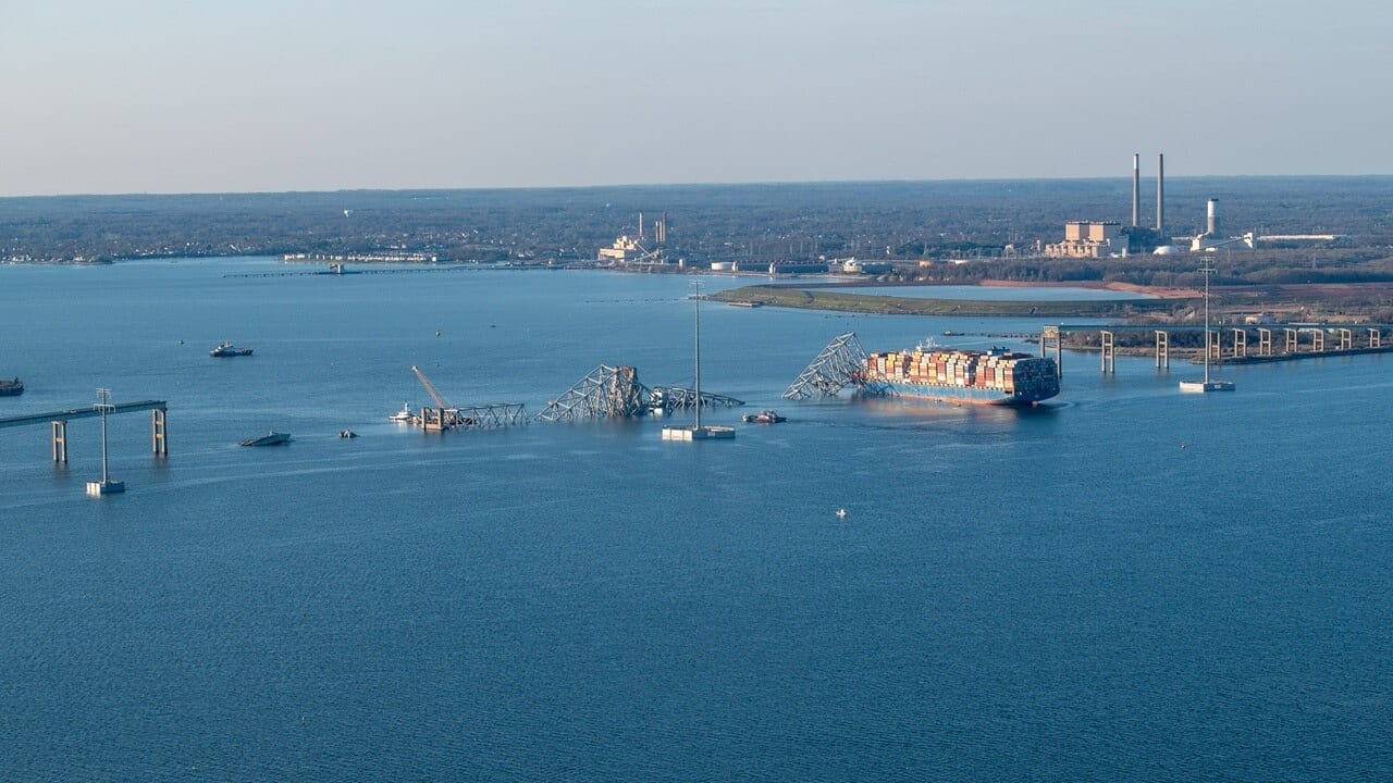 Wide aerial view of the Francis Scott Key Bridge collapse, showing the Dali stopped in the channel with the bridge’s remaining spans intact on either side of the gap.