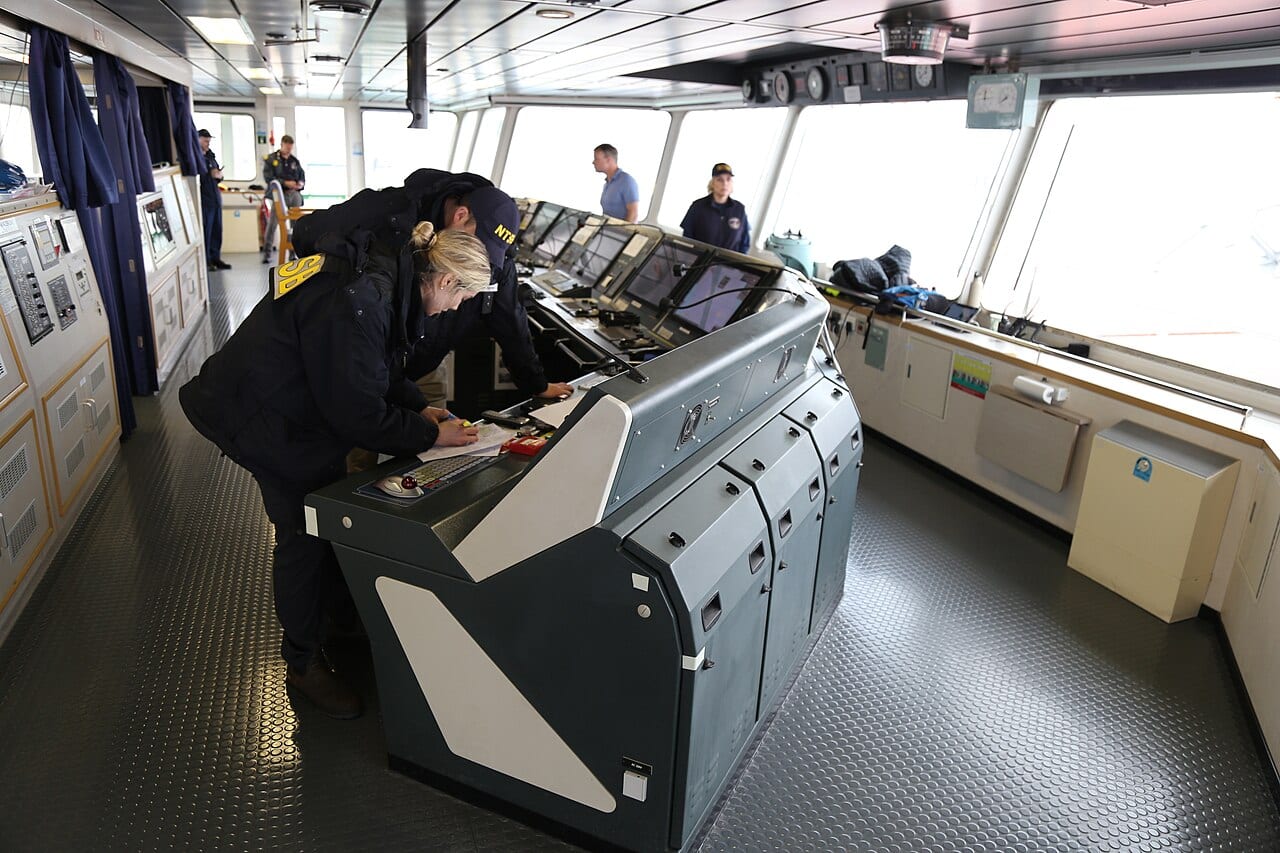 NTSB investigators examining an electrical control console on the navigation bridge of the Dali, circuit-breaker panel cabinets visible along the wall behind them.